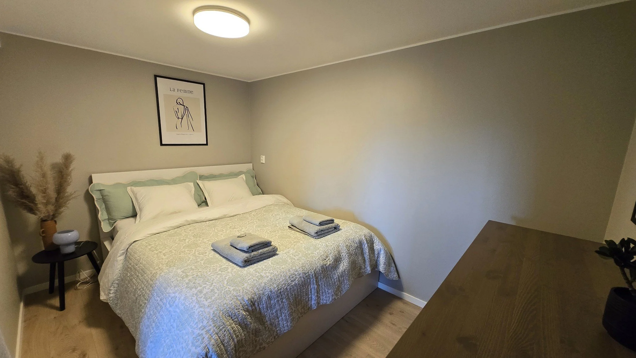 A cozy bedroom with a white bed, light green pillows, and folded towels on it. A black side table with a clay vase filled with pampas grass and a small white ceramic vase is next to the bed. A framed minimalist line drawing of a woman hangs above the