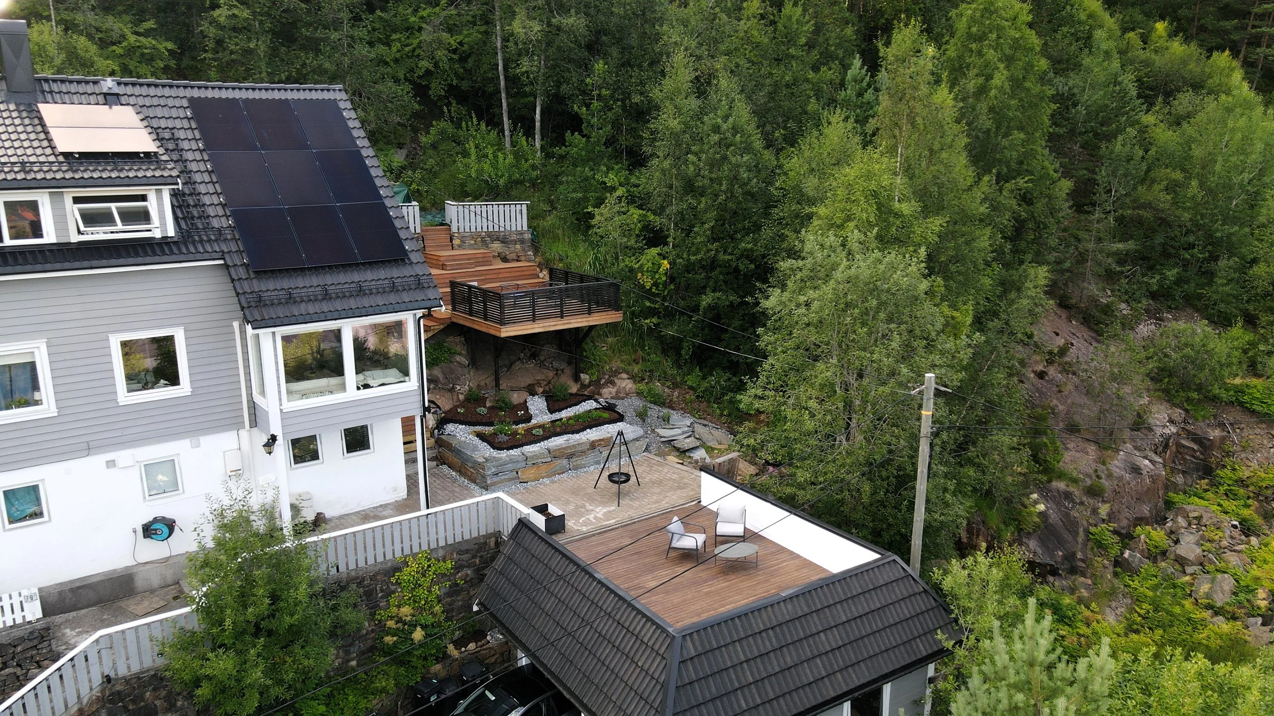 A house with solar panels on the roof, a backyard deck with outdoor furniture, a garden, and a wooded hillside in the background.
