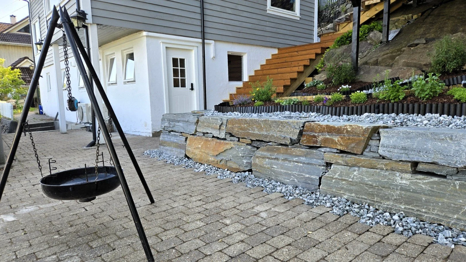 A backyard with a patio made of brick pavers, a black metal swing set, a stacked stone retaining wall, and a landscaped garden with plants and flowers. The house has gray siding, white trim, a white door, and a wooden staircase leading to an upper le