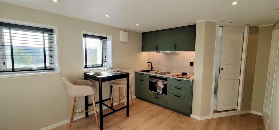 Small modern kitchen with green cabinets, a built-in oven, a stovetop, and a countertop with a faucet. Adjacent to the kitchen area is a high table with two beige chairs. Two windows with black blinds are on the wall near the table, and there is a do