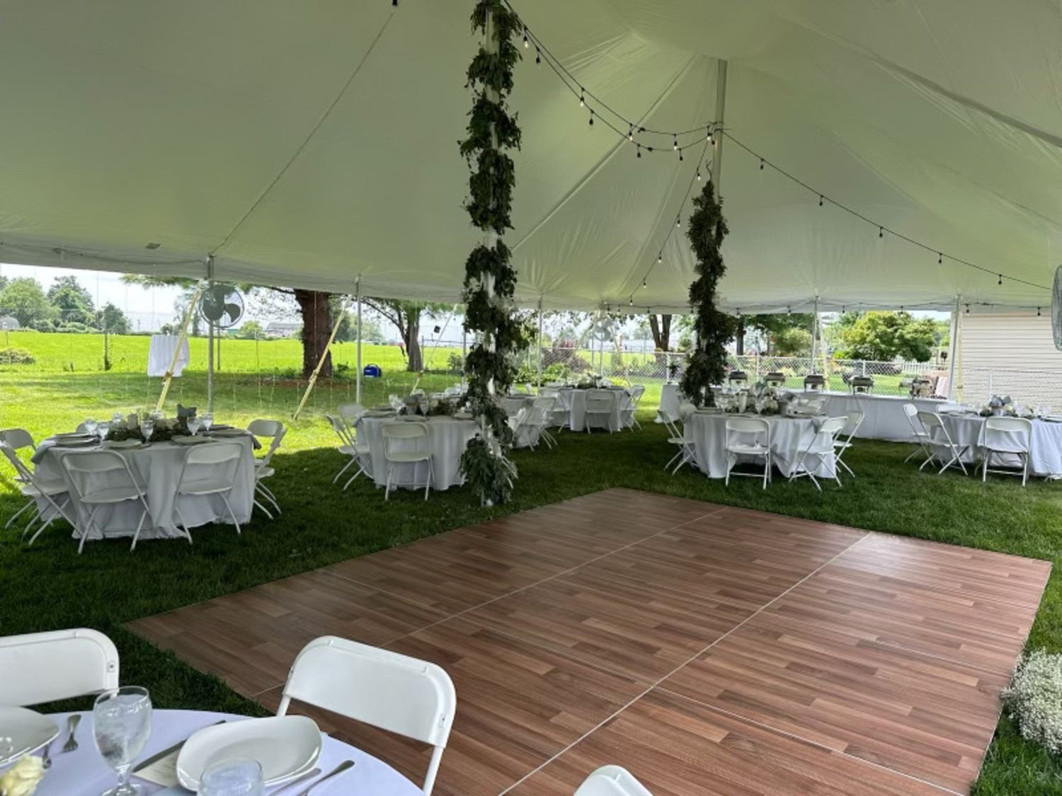 Outdoor event tent with round banquet tables covered in white tablecloths and set with plates, glasses, and napkins. A wooden dance floor area is visible, and string lights and greenery decorations hang inside the tent. Green grass and trees can be s