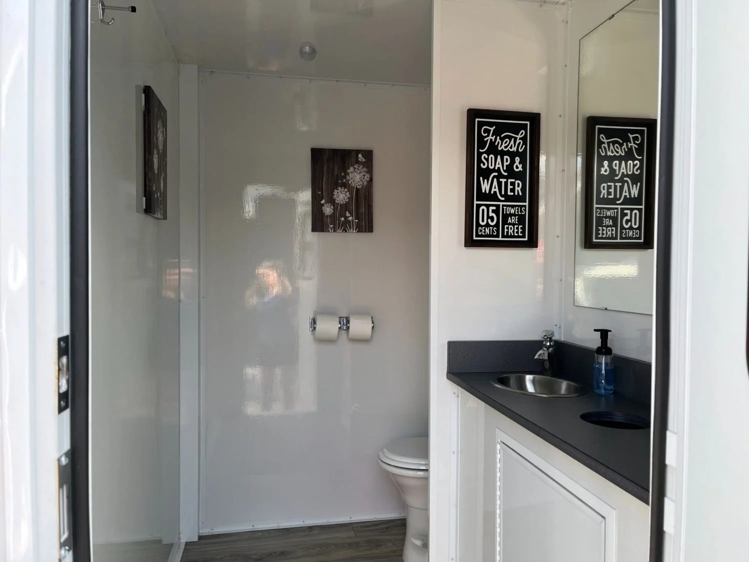 Small bathroom with white walls, a toilet, a black countertop with a stainless steel sink, a soap dispenser, and framed signs on the wall that say "Fresh Soap & Water" and "Towels Are Free."