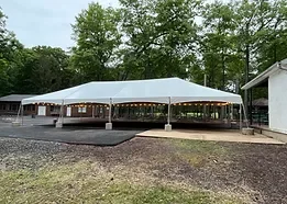 A large outdoor pavilion with a white roof, situated on a paved area with grassy surroundings and nearby trees.
