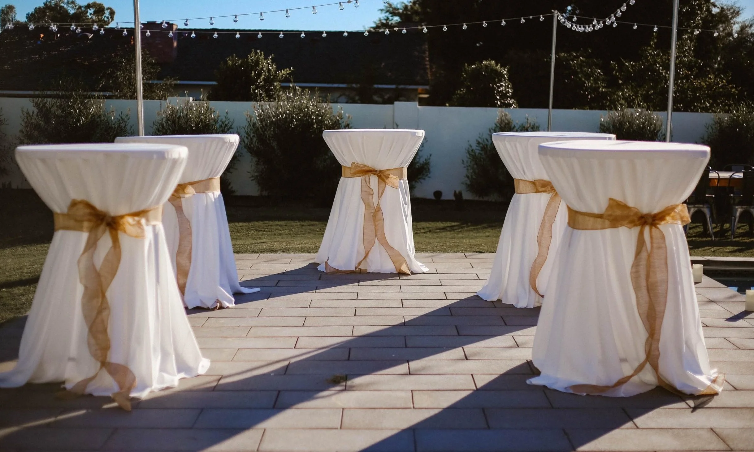 Outdoor event setup with five high cocktail tables draped in white cloth and tied with gold ribbons, on a tiled patio with string lights overhead and bushes in the background during dusk.