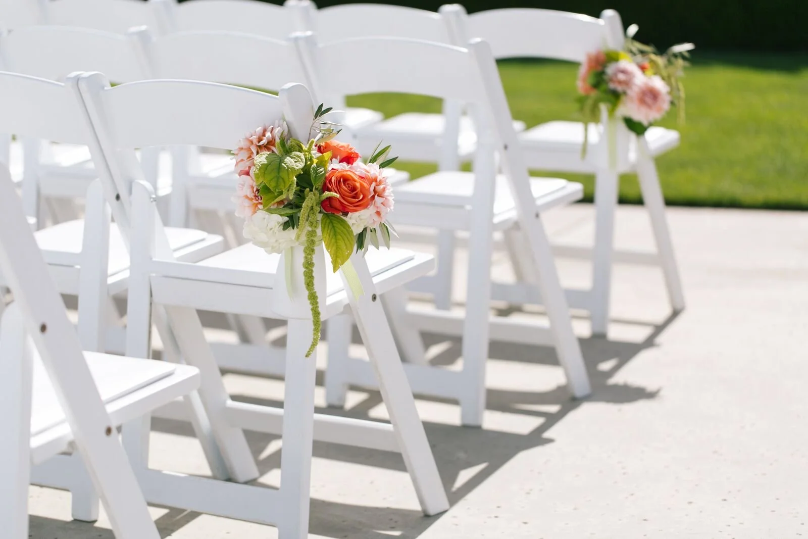 White folding chairs decorated with small bouquets of pink and orange flowers with green leaves, arranged outdoors on a paved surface with a grassy background.