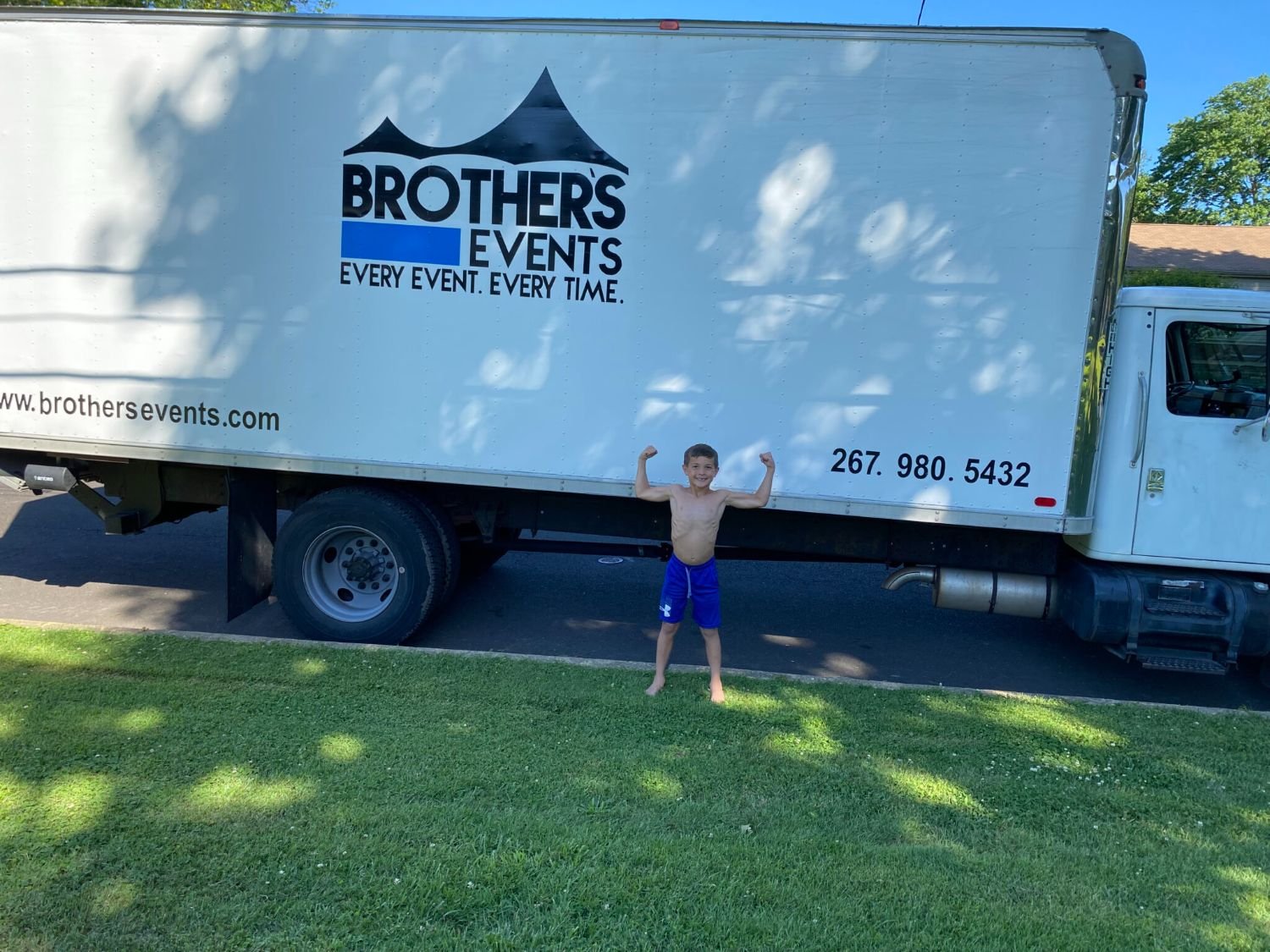 A young boy in blue shorts standing outside on grass with arms raised in a flexing pose, next to a large white moving truck with the Brothers Events logo and contact information printed on the side.