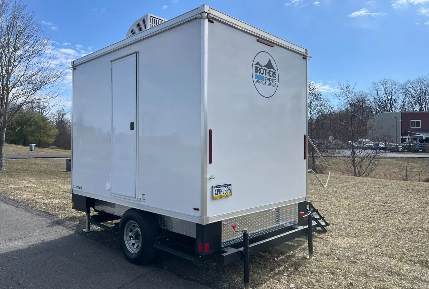 White mobile event trailer with the logo 'Brothers Events' on the side, parked on a grassy area with trees and a clear blue sky nearby.
