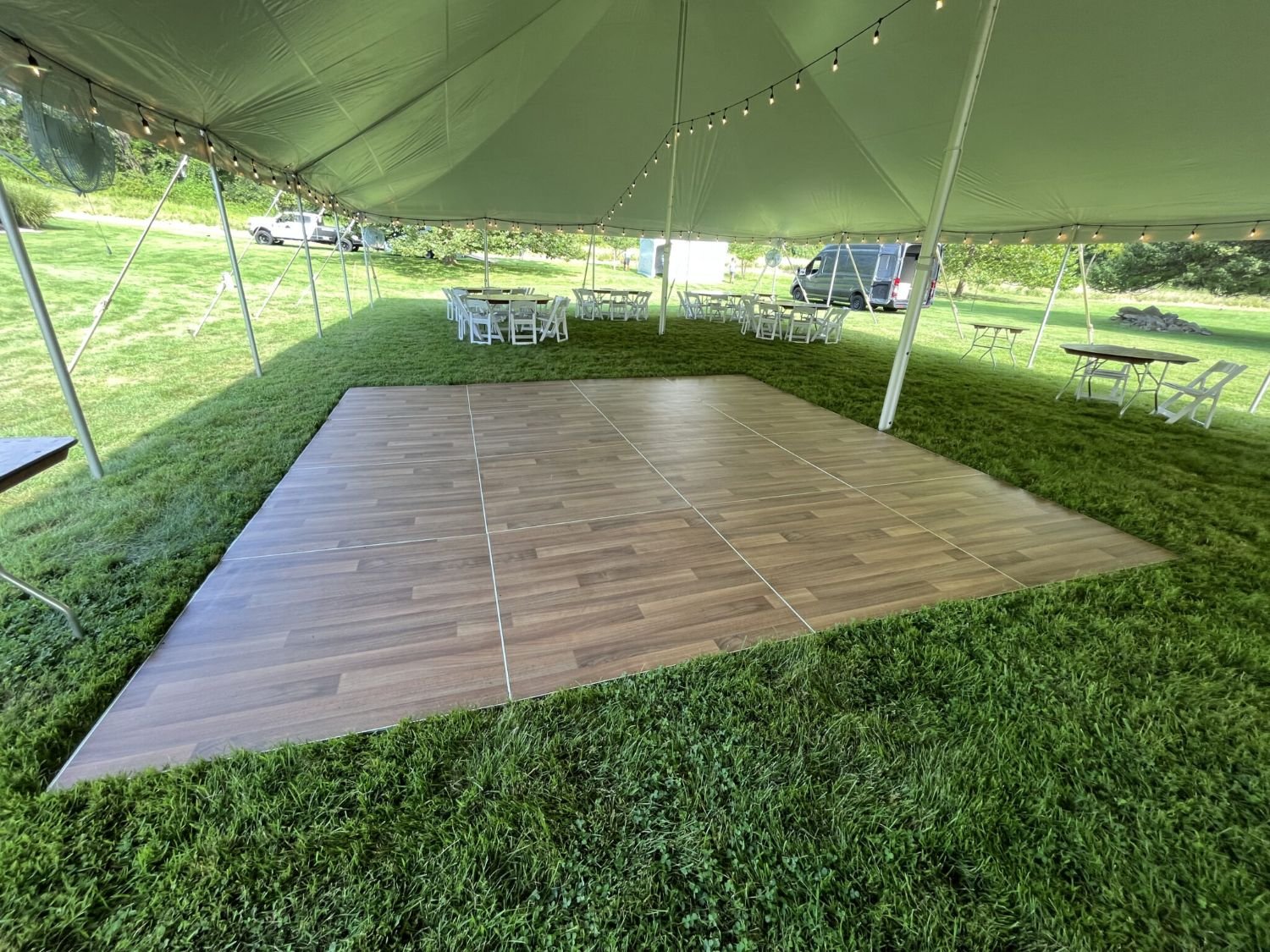 Empty wooden dance floor under a large outdoor tent with string lights, surrounded by tables and chairs on green grass.