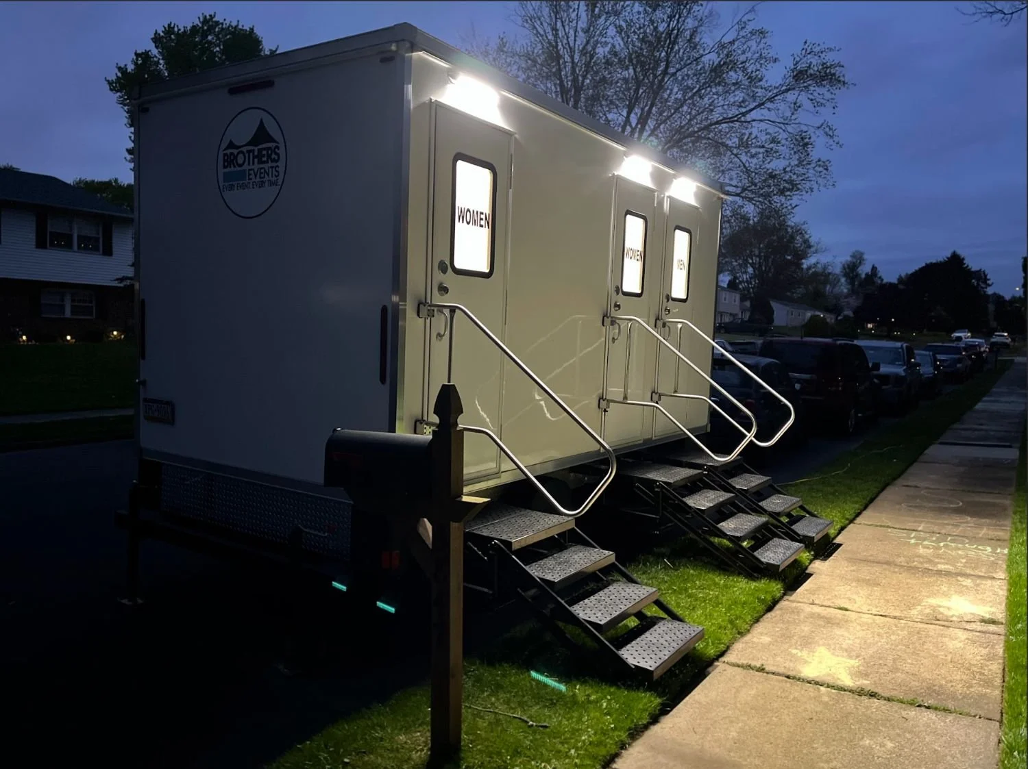 A mobile restroom trailer with women's and men's signs, located on a residential street in the evening, with stairs and handrails leading up to the doors, and parked cars along the street.