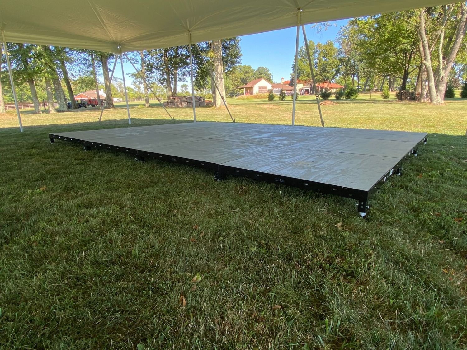 Empty outdoor stage with a beige canopy, set up on grass in a park-like area with trees and houses in the background.