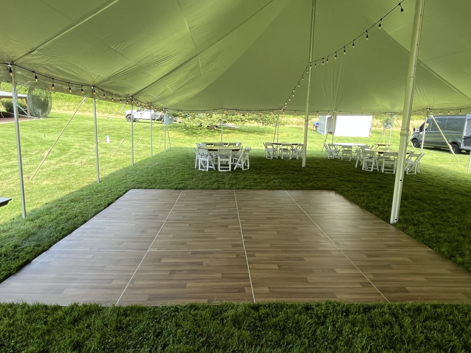 A large outdoor tent with a wooden dance floor at the front, surrounded by green grass. Inside the tent, there are several round tables with white chairs and string lights hanging from the ceiling. In the background, there are vehicles and a trailer.
