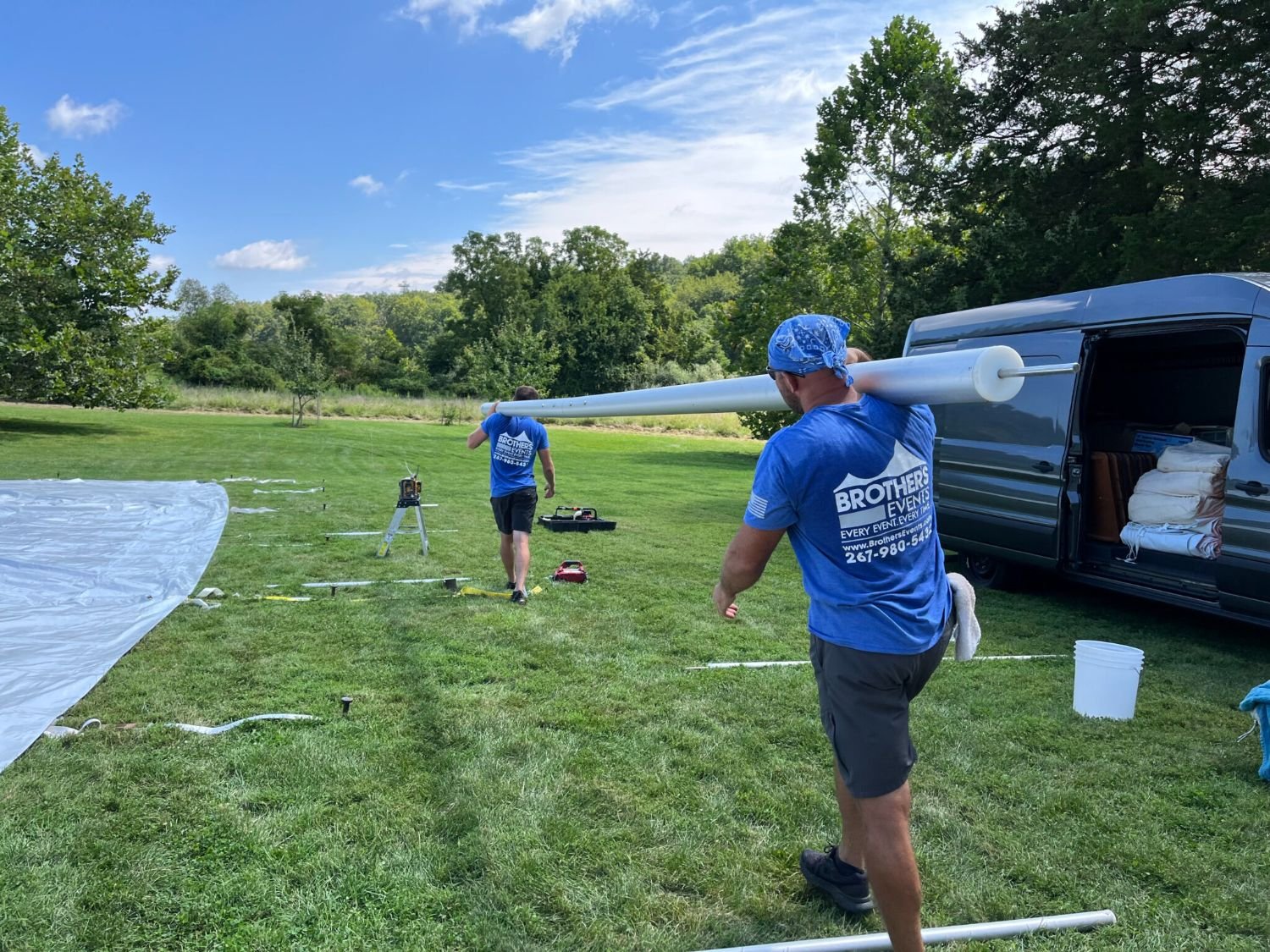 Two men in blue t-shirts working outdoors with large white tubes and equipment on a grassy field, with a van and trees in the background.