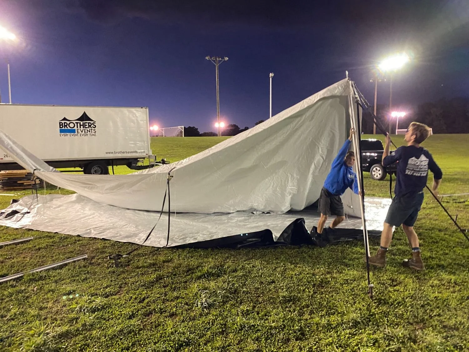 Two men are setting up a white large event tent on a grassy field at night with stadium lights in the background.