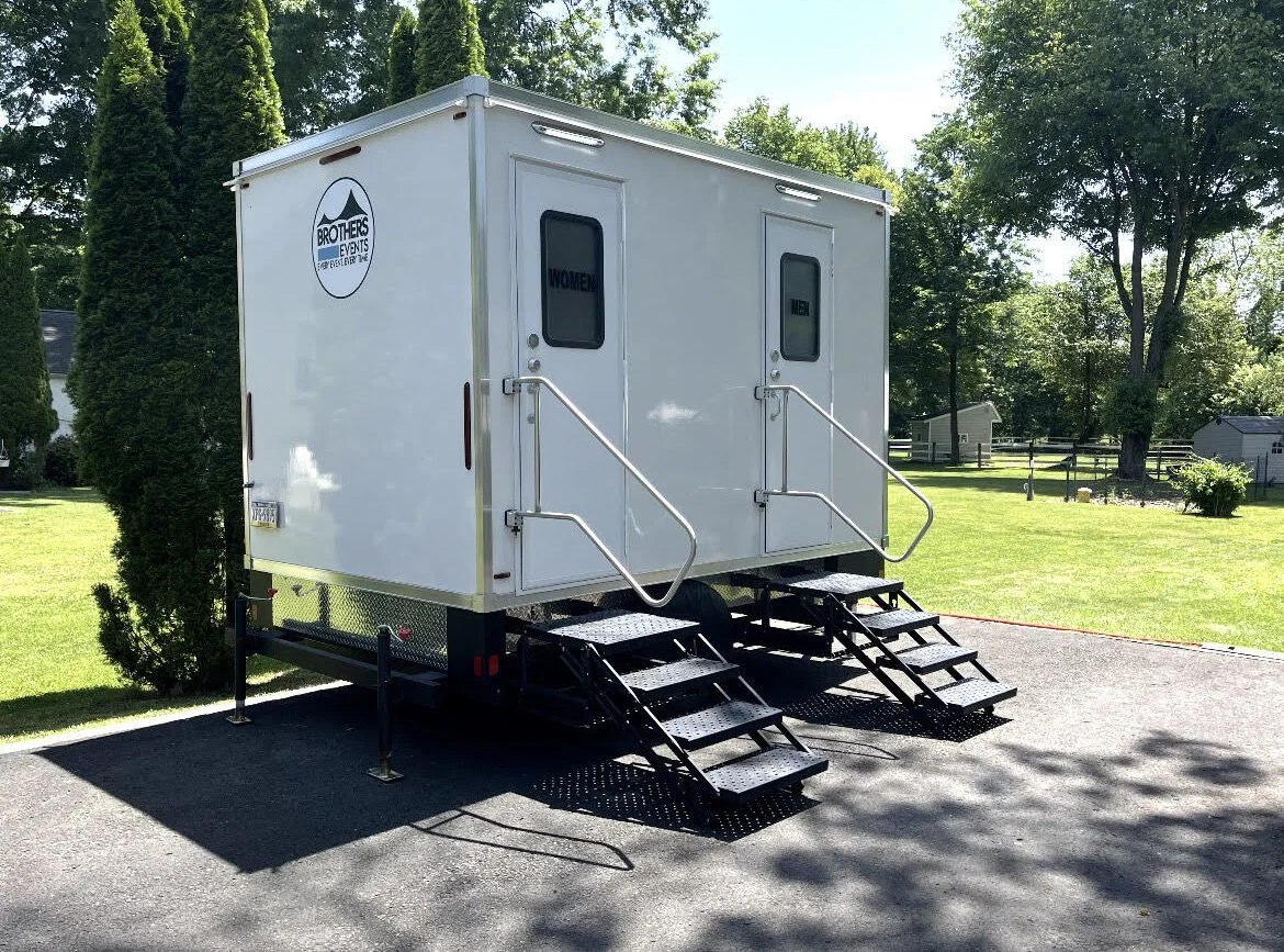 Portable restroom trailer with two doors labeled 'Men' and 'Women', set up outdoors on a paved surface in a grassy area with trees.