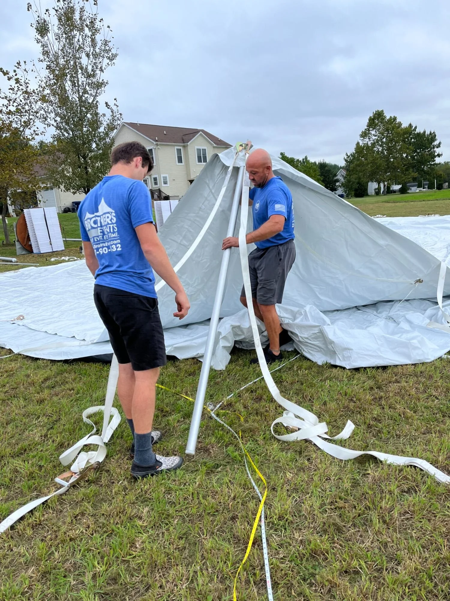 Two men are setting up a large tent on a grassy area, with house and trees in the background. They are wearing matching blue shirts and handling tent poles and fabric.
