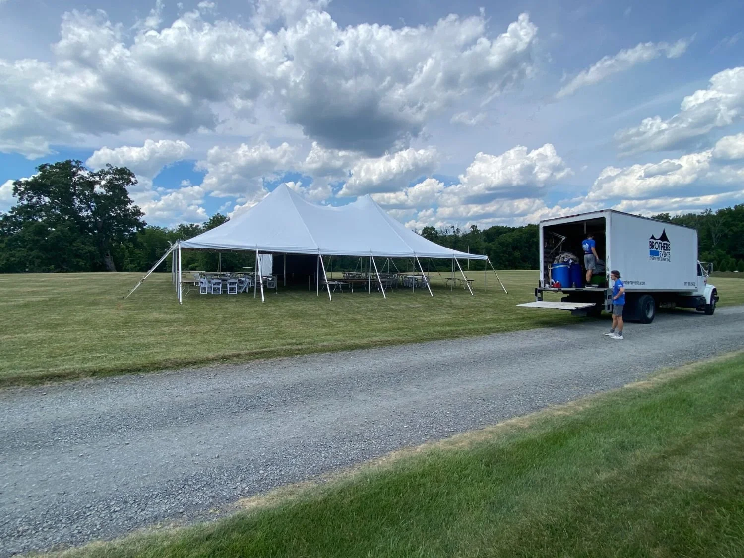 Setup for an outdoor event with a large white tent and a mobile food truck on a grassy field under cloudy skies.