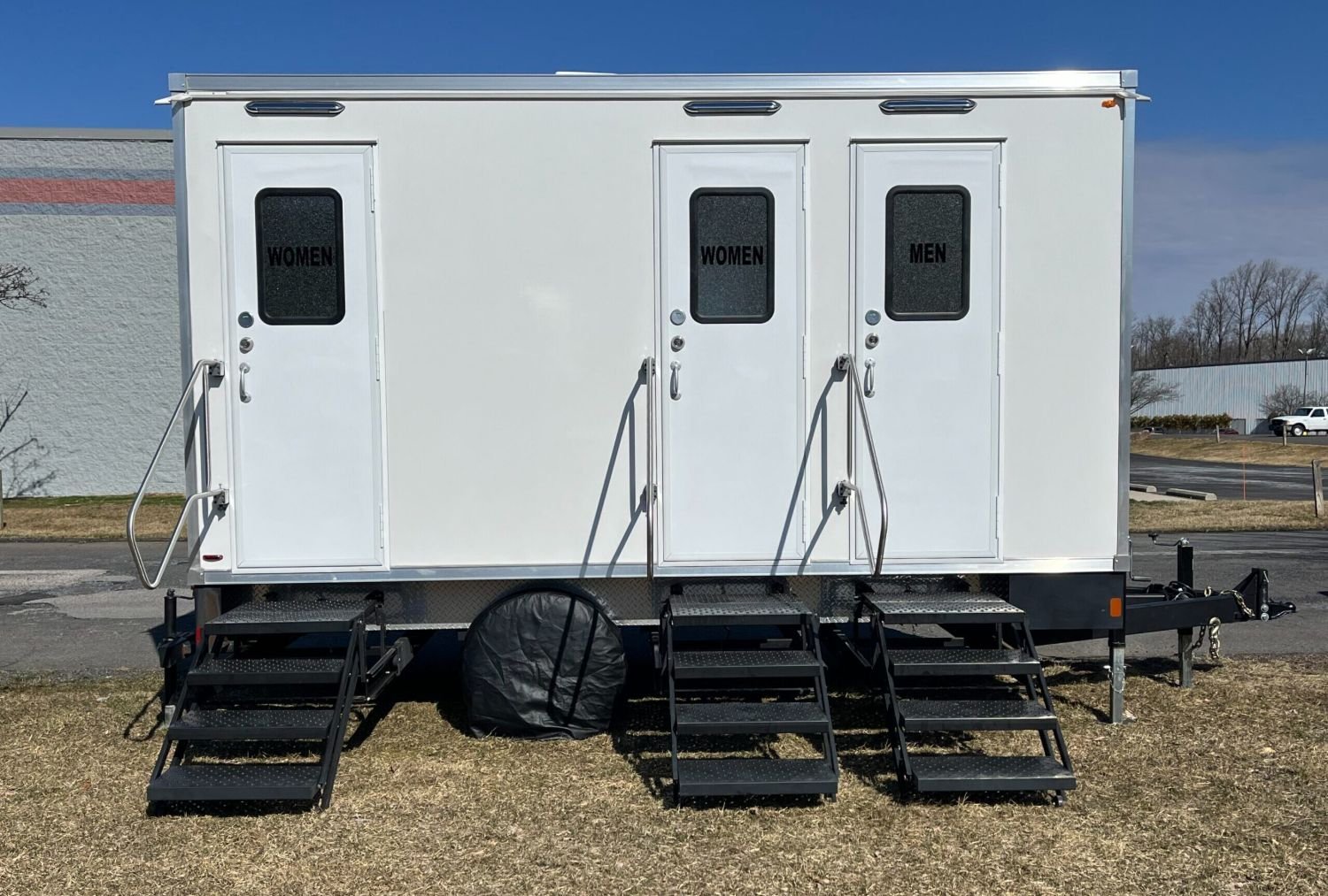 Mobile restroom trailer with separate entrances for women and men, each with a small window, metal stairs, and handrails, parked on grass with trees and buildings in the background.