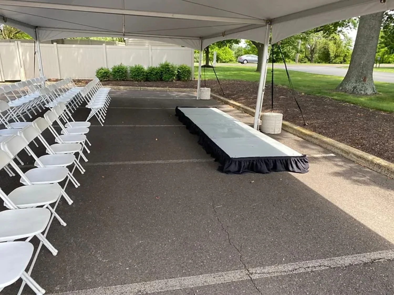 Empty outdoor event setup with white chairs arranged in rows, a stage with a gray top and black skirt, under a white canopy tent on an asphalt lot, with a grassy area, trees, and a car in the background.