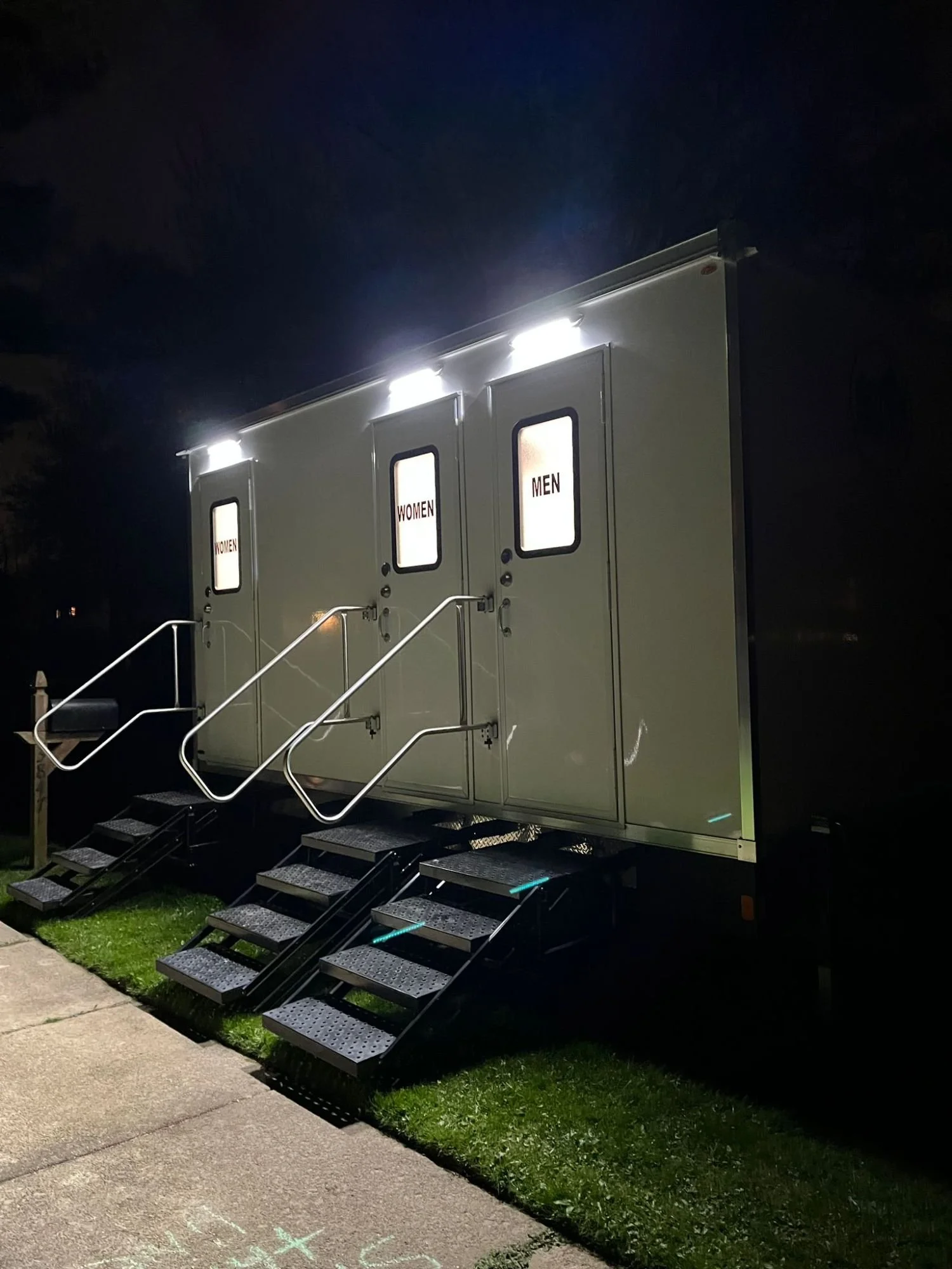 A portable restroom with separate entrances labeled WOMEN and MEN, illuminated at night.