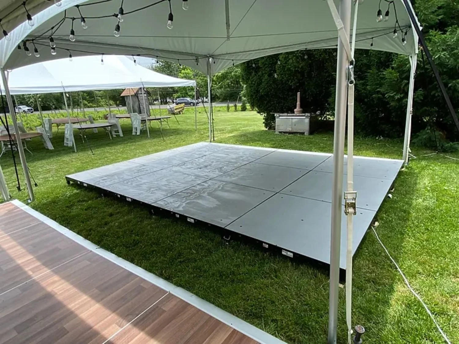Empty event tent with a raised dance floor and string lights in a grassy outdoor area, with tables and chairs visible in the background.