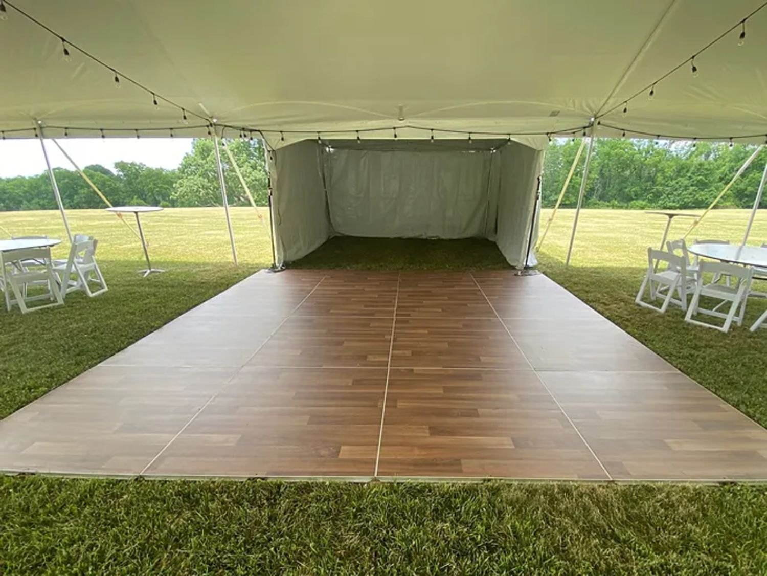 Empty event tent with a wooden dance floor, white tables and chairs on a grassy field, surrounded by trees.