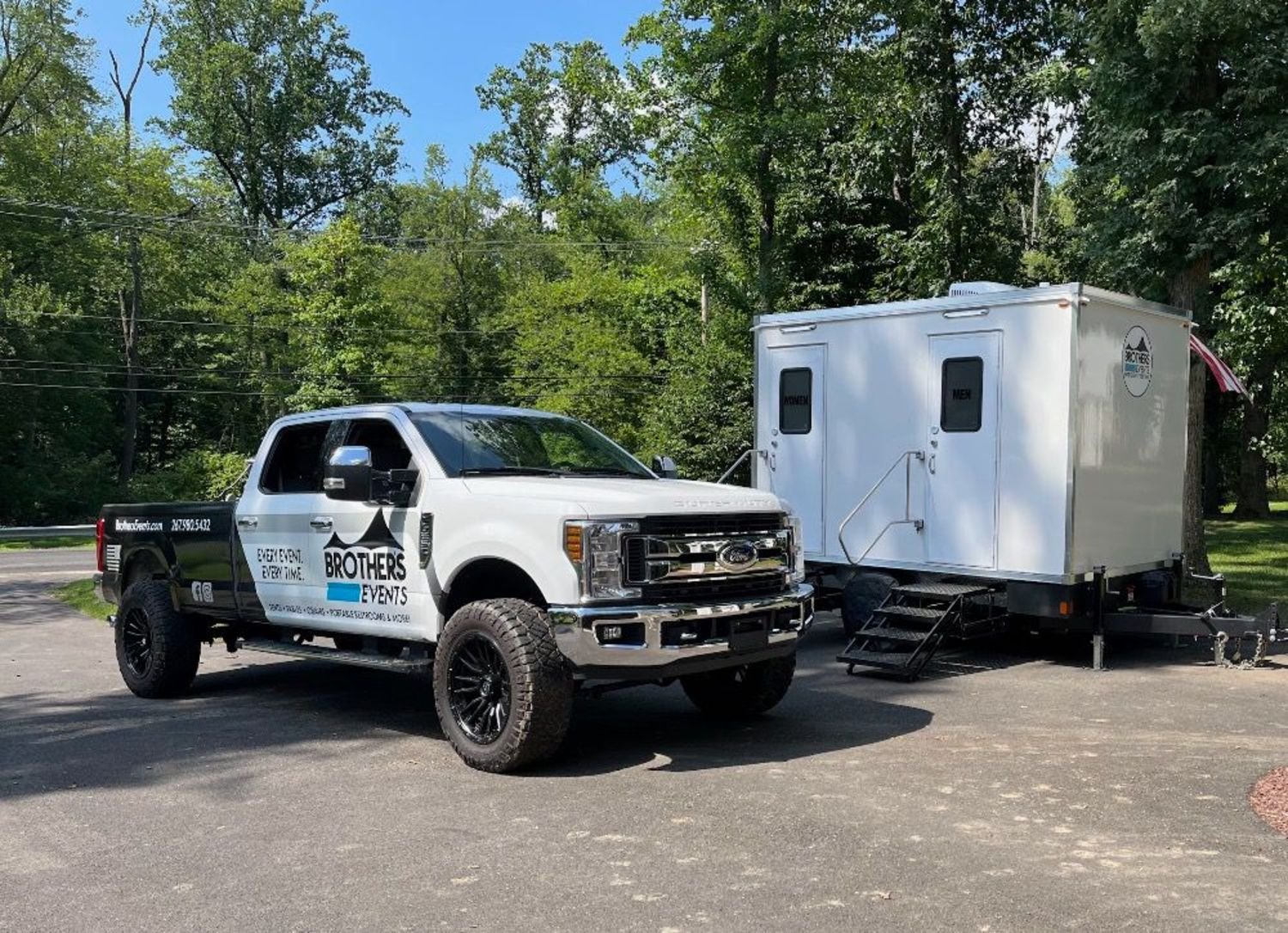 A white pickup truck with advertising for Brothers Events parked on a paved surface. Next to it is a white portable restroom trailer with two doors labeled 'Women' and 'Men', set against a backdrop of green trees and a blue sky.