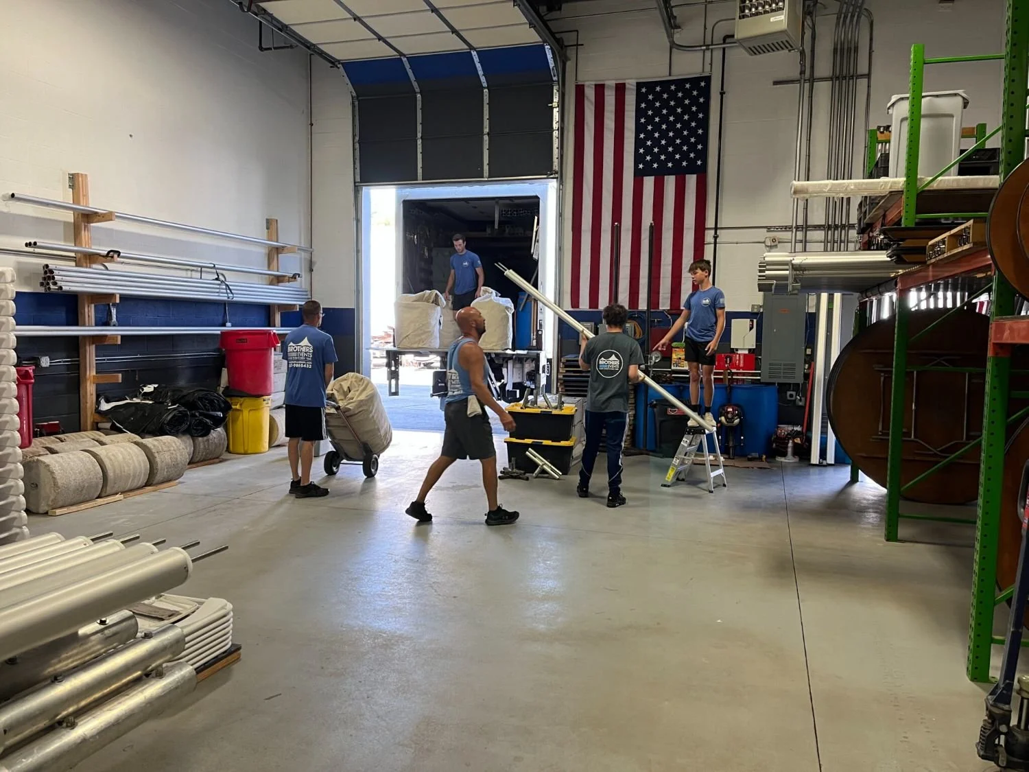 Group of five young men unloading large white bags from a truck in a warehouse with American flag on the wall.