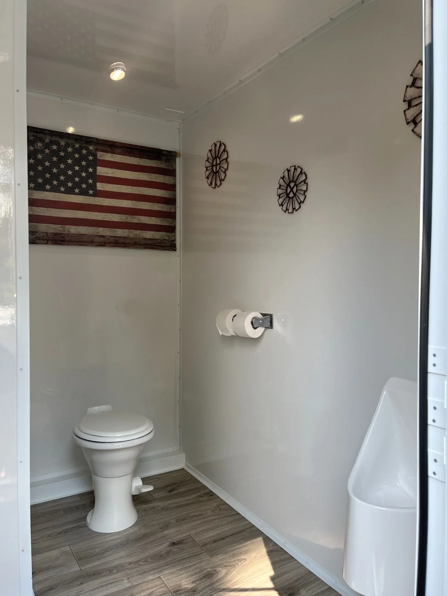 A small bathroom with a white toilet, a toilet paper holder, a urinal, a wall decoration, and an American flag poster on the wall.