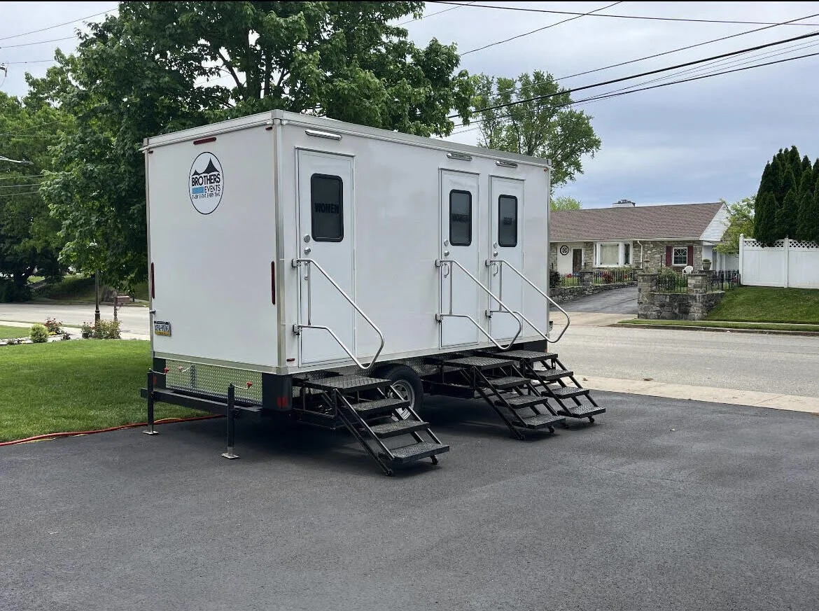 A mobile event trailer with steps and railings parked on a paved driveway, with a house, trees, and power lines in the background.