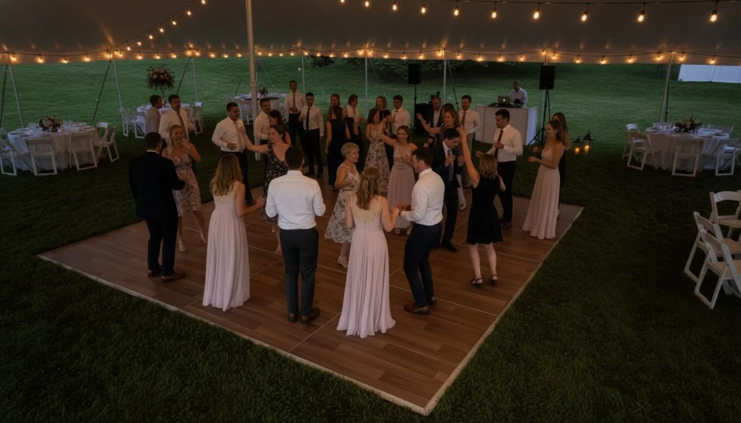 People dancing on a wooden dance floor at an outdoor evening wedding reception under a tent with string lights, surrounded by tables with white chairs and floral centerpieces.
