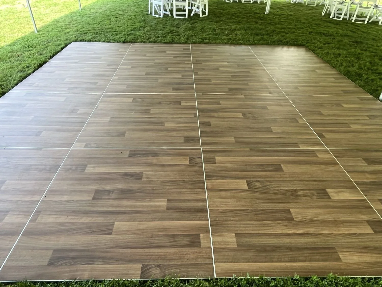 Wooden dance floor set up on grass at outdoor event with white chairs in background.