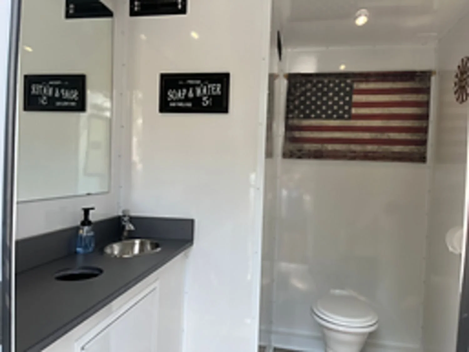 Small bathroom with a black countertop sink, soap dispenser, mirror, and signs on the wall; American flag decoration on the wall above the toilet.