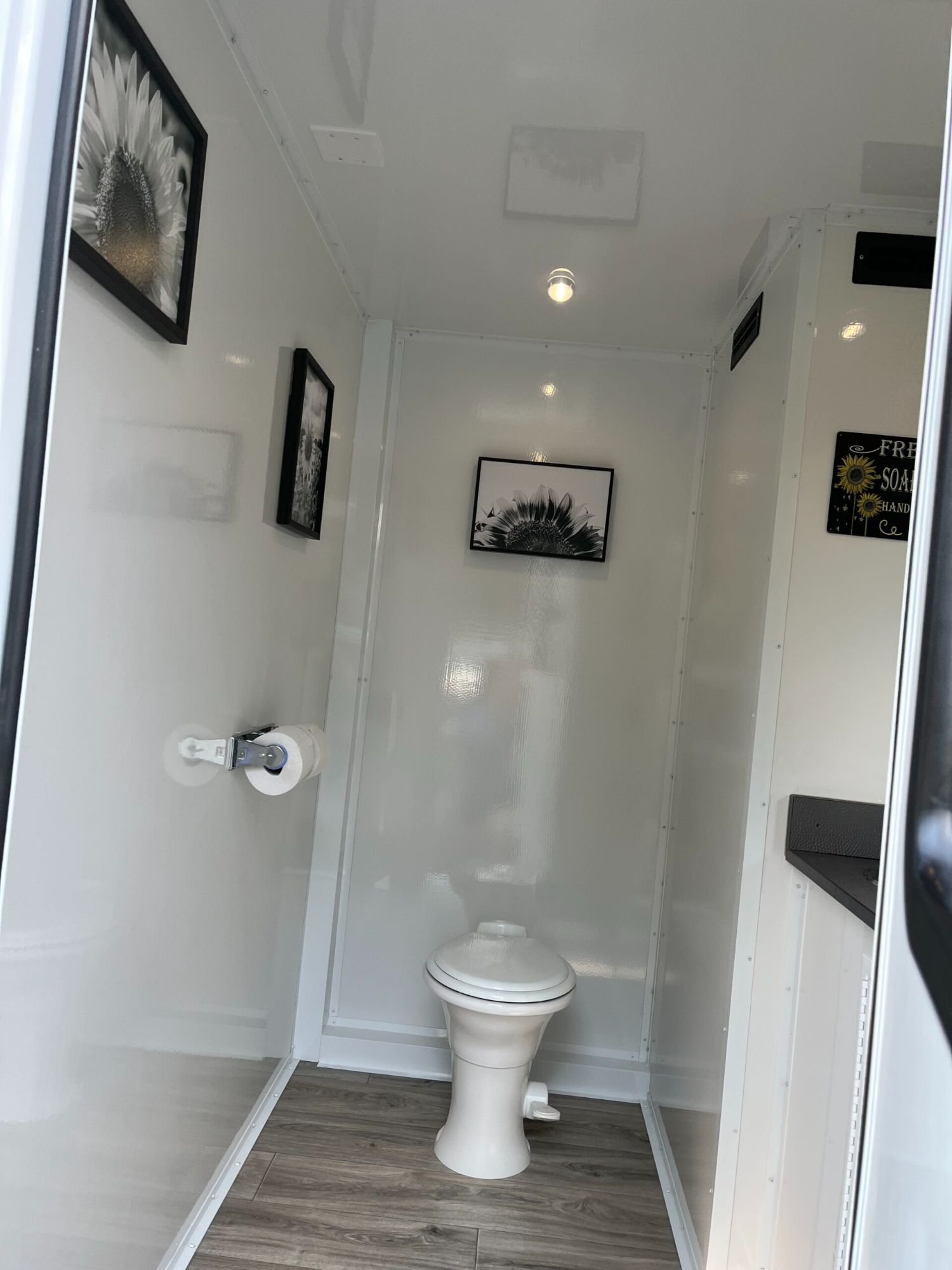 Small restroom with white walls, a wooden floor, framed black and white flower artwork, a toilet, and a roll of toilet paper on a holder.
