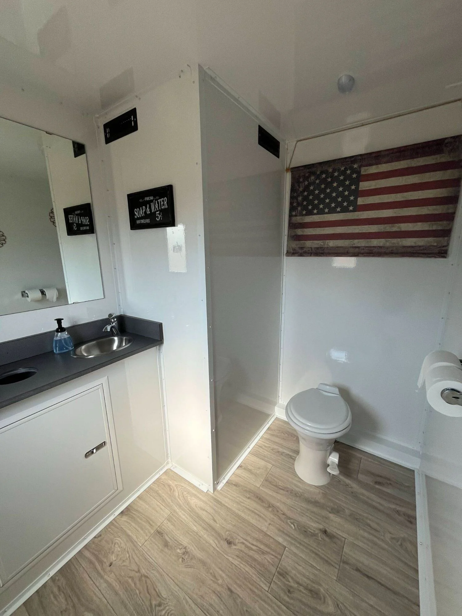 A small bathroom with a white vanity, a mirror, a soap dispenser, a toilet, and an American flag-themed curtain.