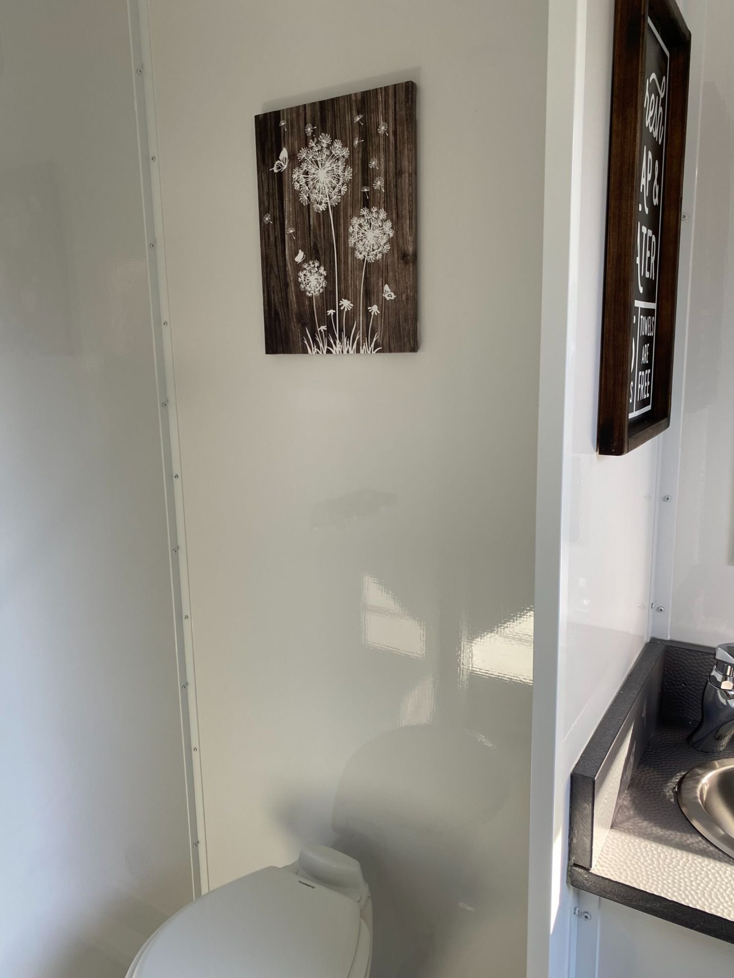 A small bathroom with a white toilet, a decorative wooden wall art with white flowers and butterflies, and a partial view of a black countertop with a stainless steel sink.