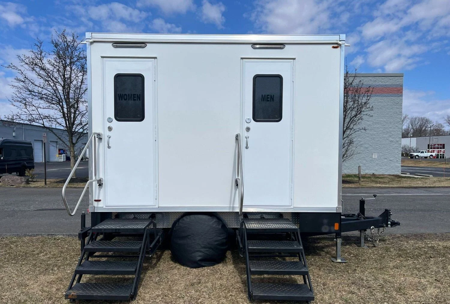 White portable restroom trailer with two separate doors labeled women's on the left and men's on the right, with metal stairs and handrails outside each door, parked on grass and asphalt under a partly cloudy sky.
