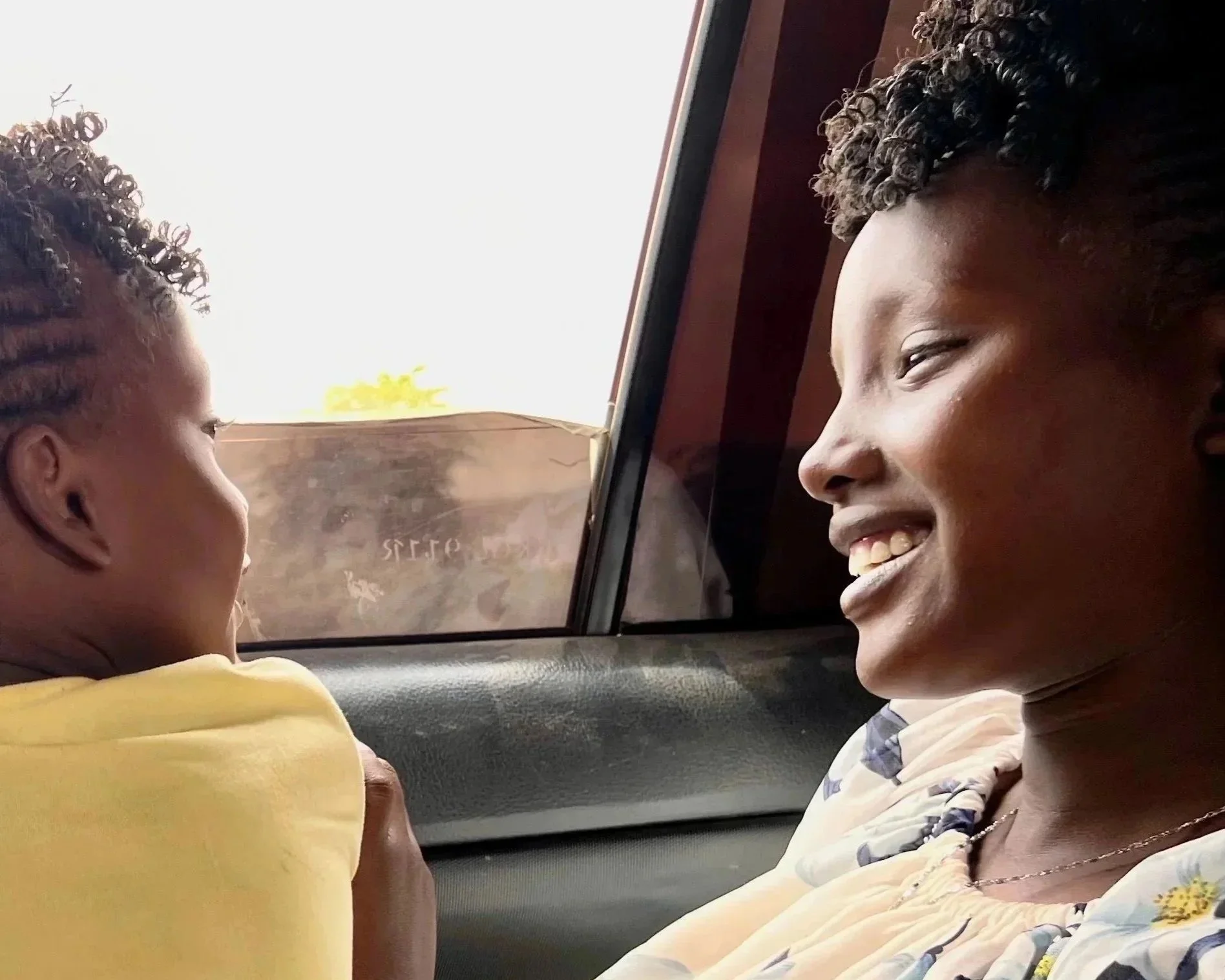 A woman smiling and holding a young child in a car with a window showing a bright sky outside.