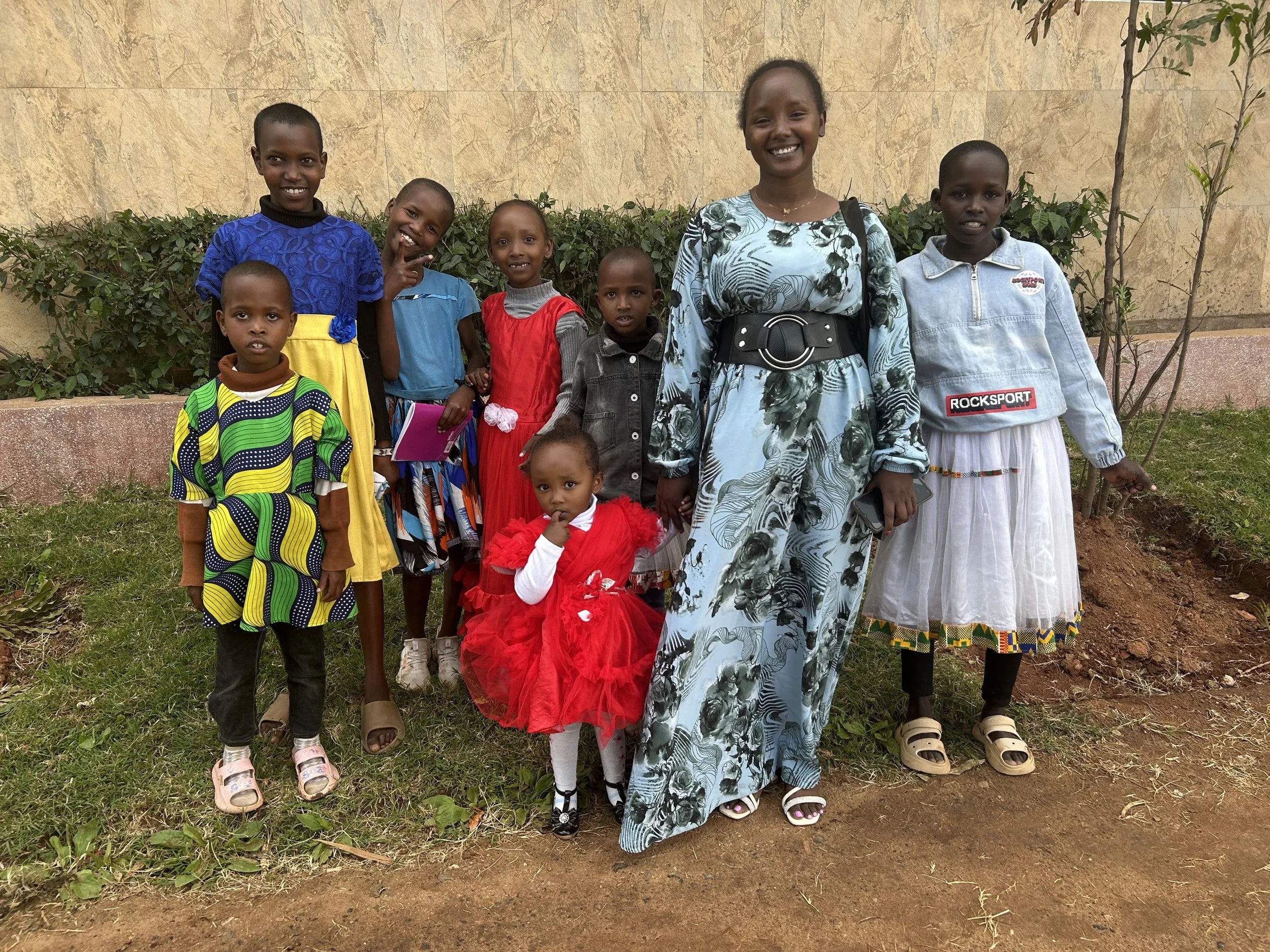 A group of nine children and one woman standing outdoors on grass, smiling for the camera. They are dressed in colorful clothing, with some children wearing dresses and others in casual outfits. The background features a beige wall, some greenery, an