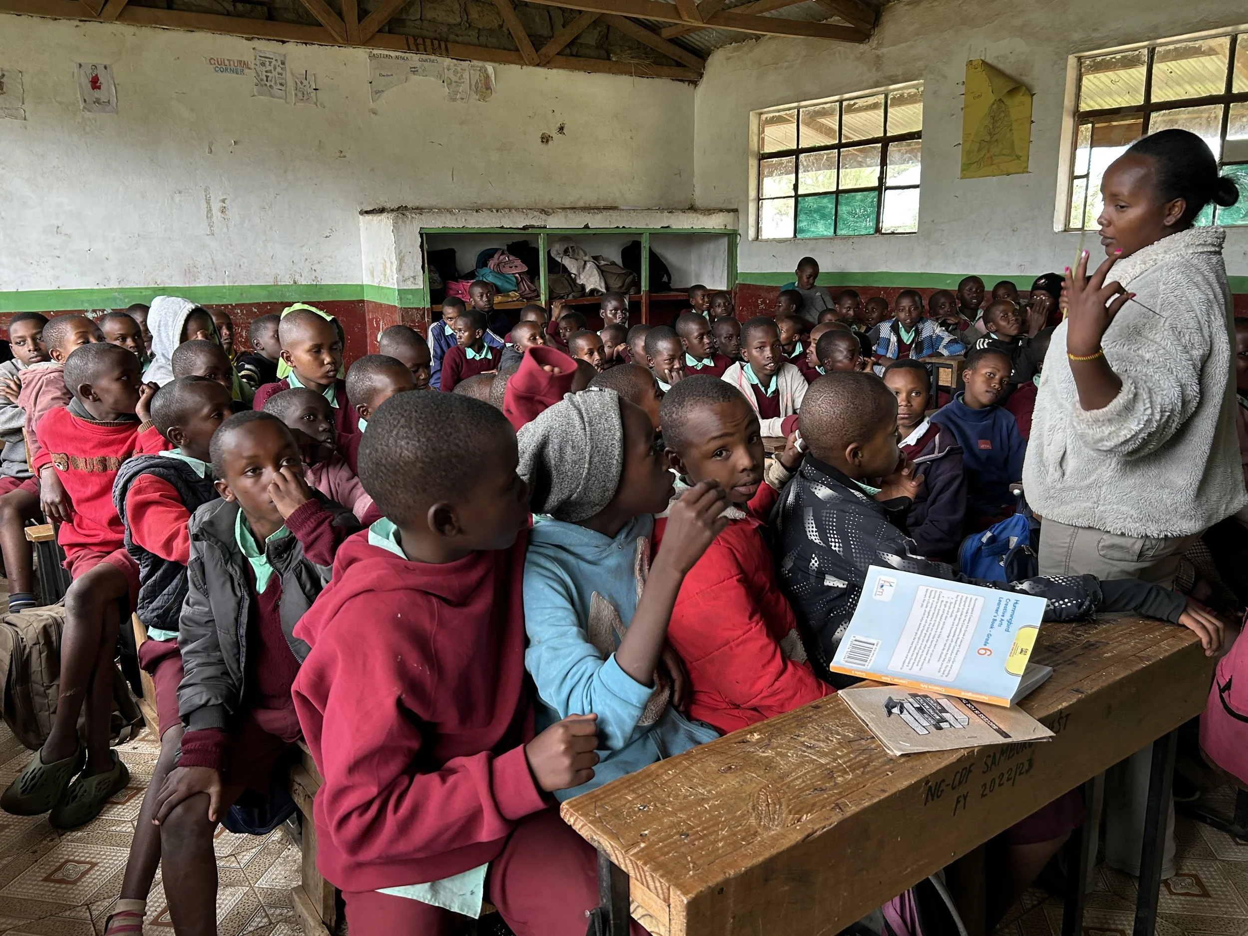 A classroom filled with young students of African descent, seated at desks and listening to a female teacher standing at the front, gesturing with her hand.