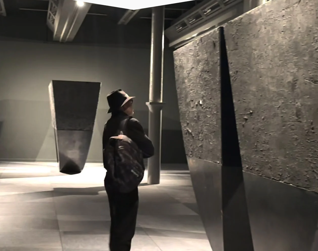 A person wearing a black hat and backpack standing in an art gallery, observing large stone and metal sculptures.