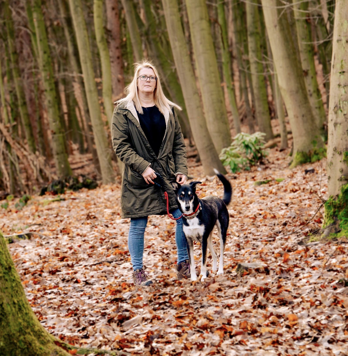 A woman with blonde hair, glasses, wearing a green jacket, black shirt, blue jeans, and hiking shoes, standing in a forest with a black and white dog on a red leash during autumn.
