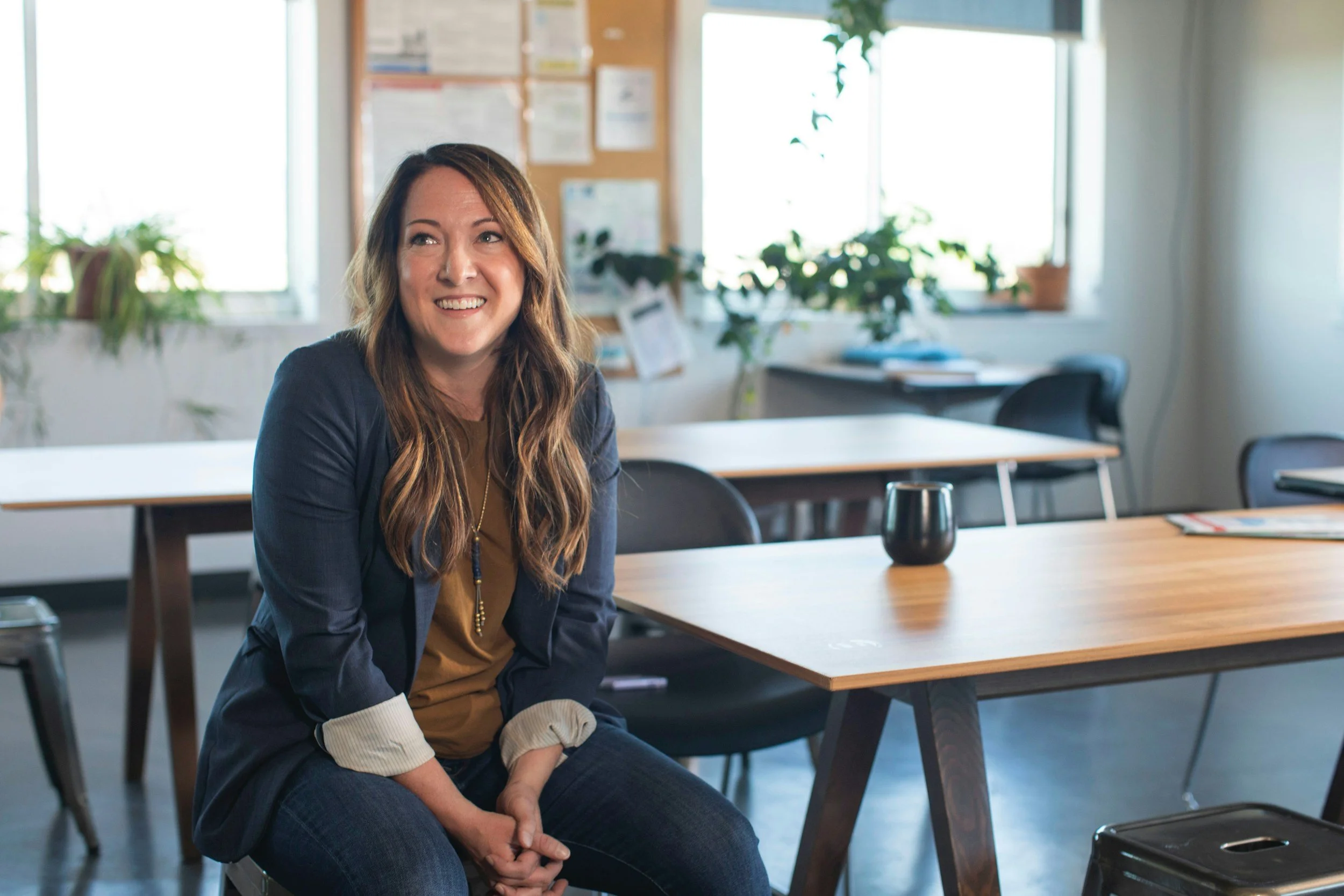A woman with long wavy hair sitting in a bright, modern office space with wooden desks, black chairs, green plants, and large windows in the background.