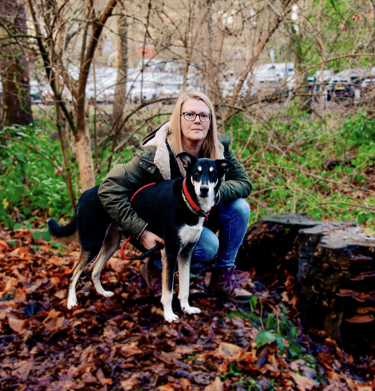 A woman with glasses and a green jacket crouches next to a black and white dog in a wooded area with fallen leaves.
