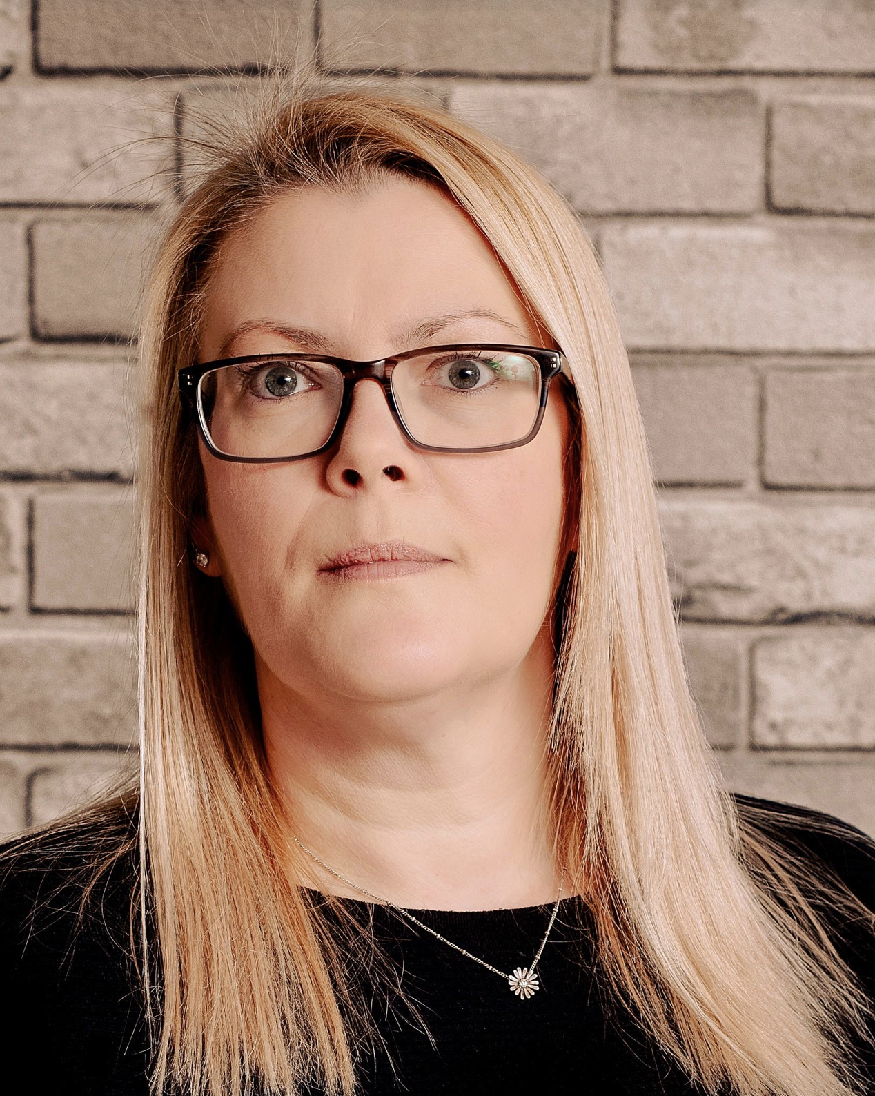 A woman with blonde hair, wearing black-rimmed glasses and a black top, standing in front of a gray brick wall.