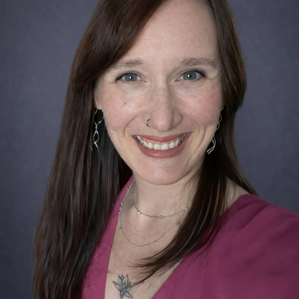 A woman with long brown hair, blue eyes, wearing a pink top, earrings, a nose ring, and layered necklaces, smiling at the camera against a plain background.