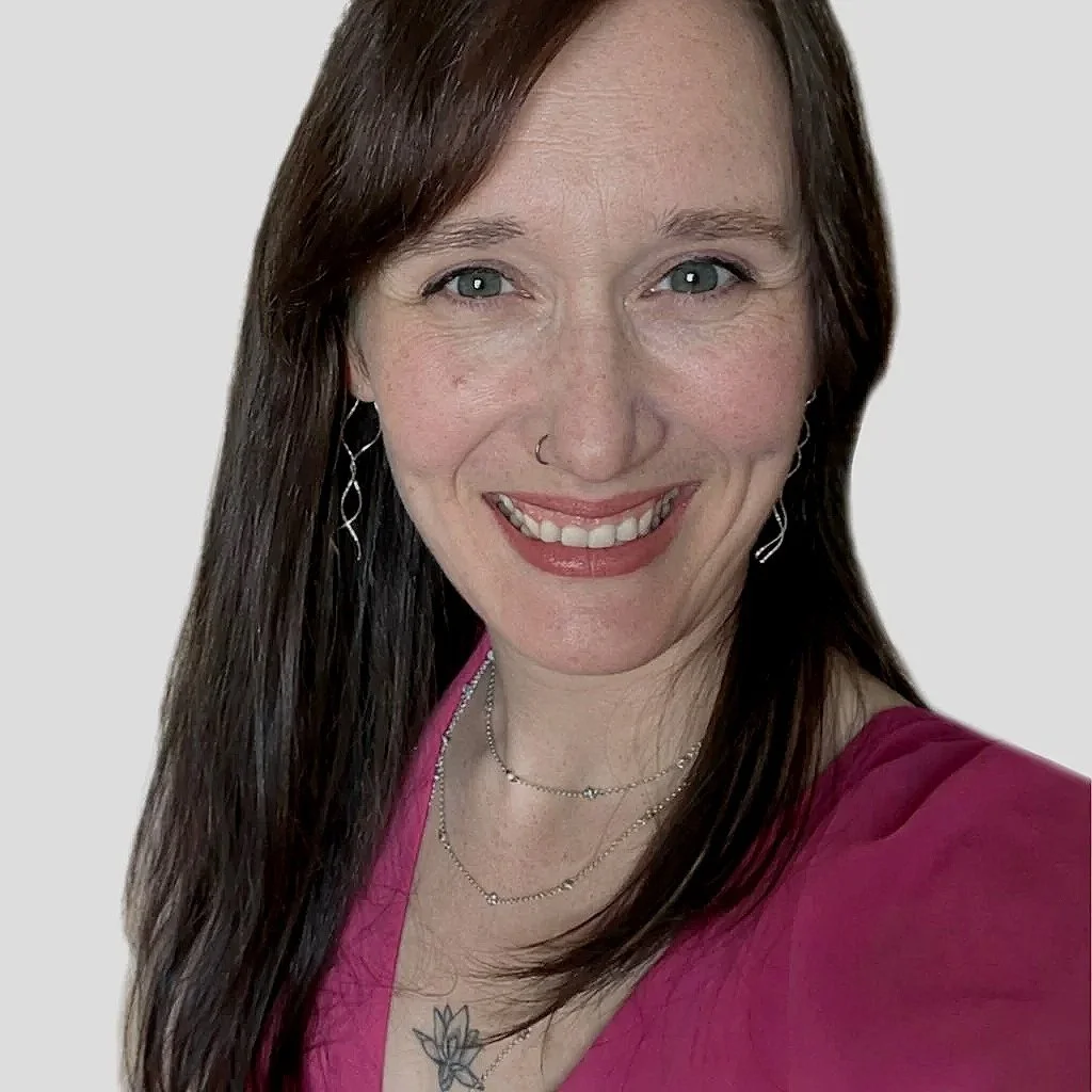 A woman with long brown hair, blue eyes, wearing a pink top, earrings, a nose ring, and layered necklaces, smiling at the camera against a plain background.