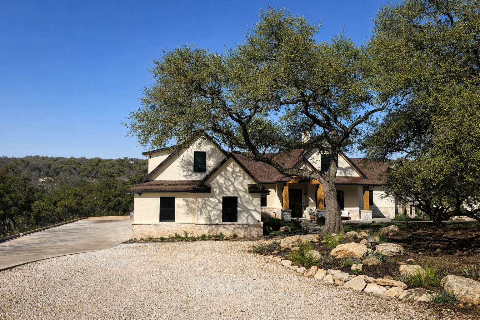 Modern house with stone and stucco exterior, large front yard with gravel driveway, mature trees, and landscaped garden.
