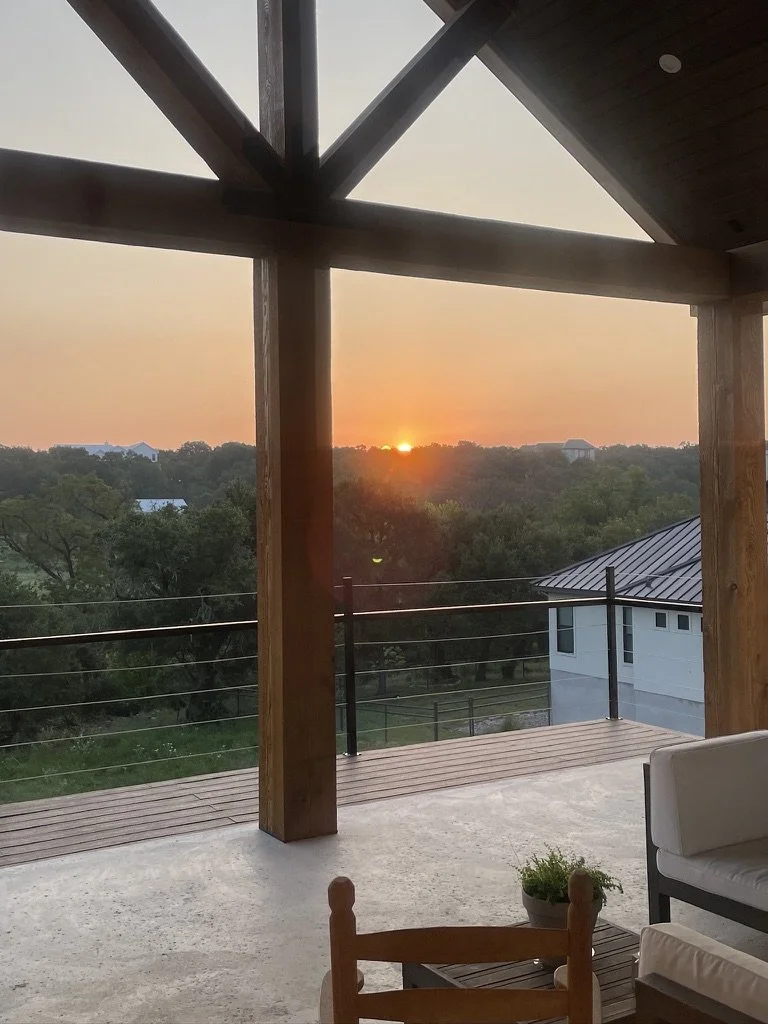Sunset view from a balcony with wooden beams, a glass railing, and furniture, overlooking trees and houses.