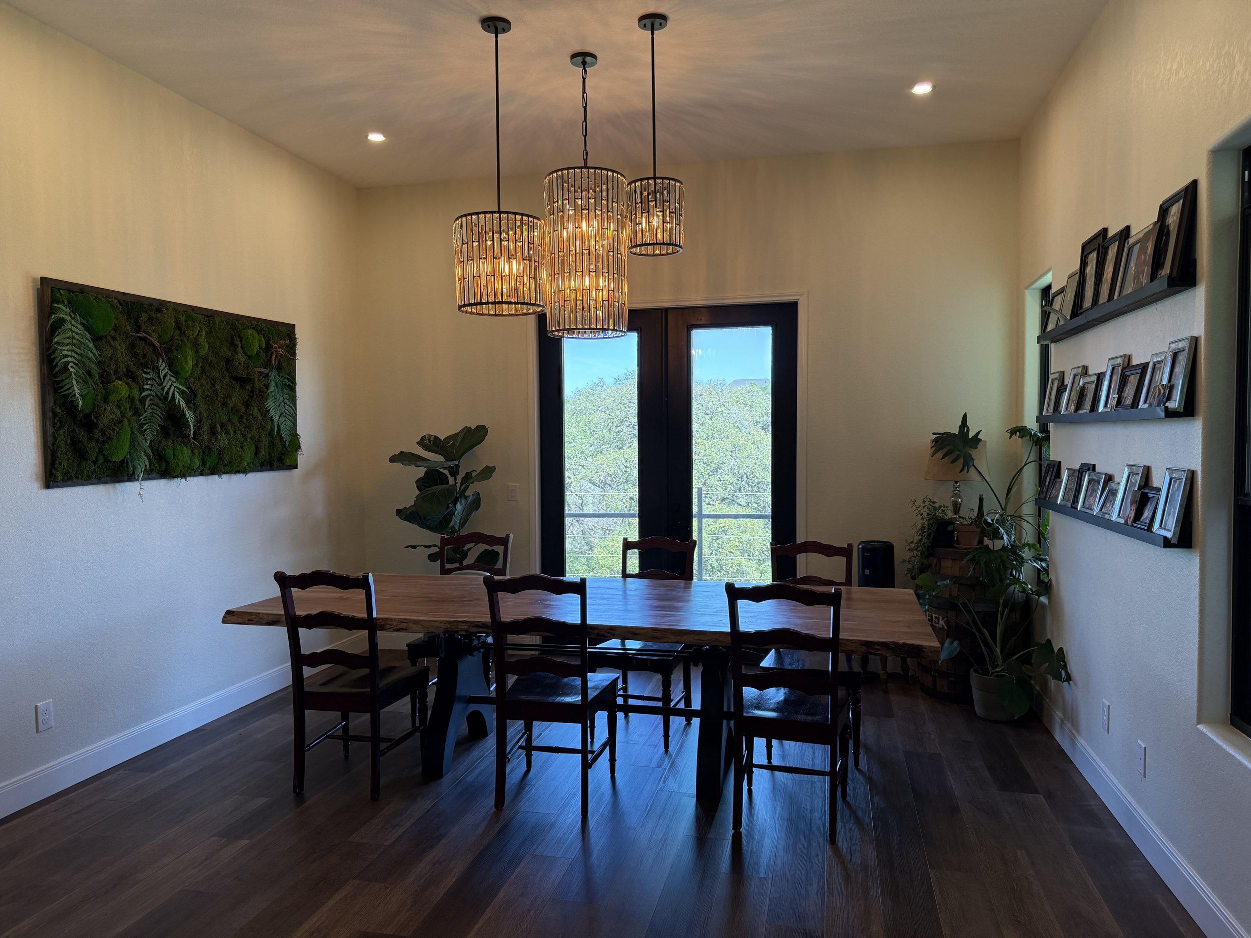 A dining room in a custom built home by Capricorn Concepts with a wooden table and six chairs, hanging ceiling lights, a large window with a balcony, potted plants, wall art with framed photos, and hardwood floors.