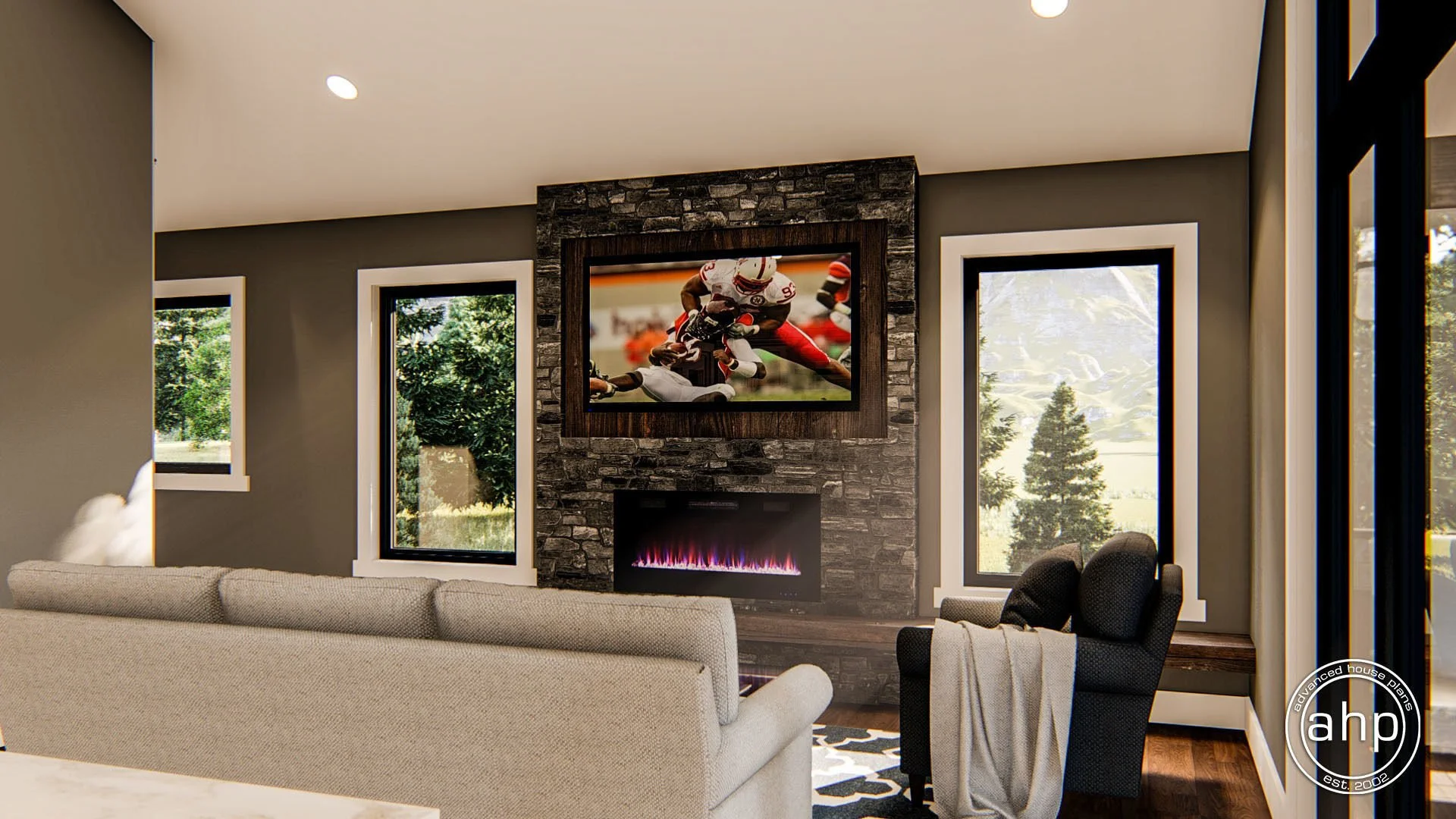 Living room with a stone fireplace, a mounted flat-screen TV displaying a football game, two large windows showing green trees outside, a beige sofa, and a black armchair with a blanket draped over it.